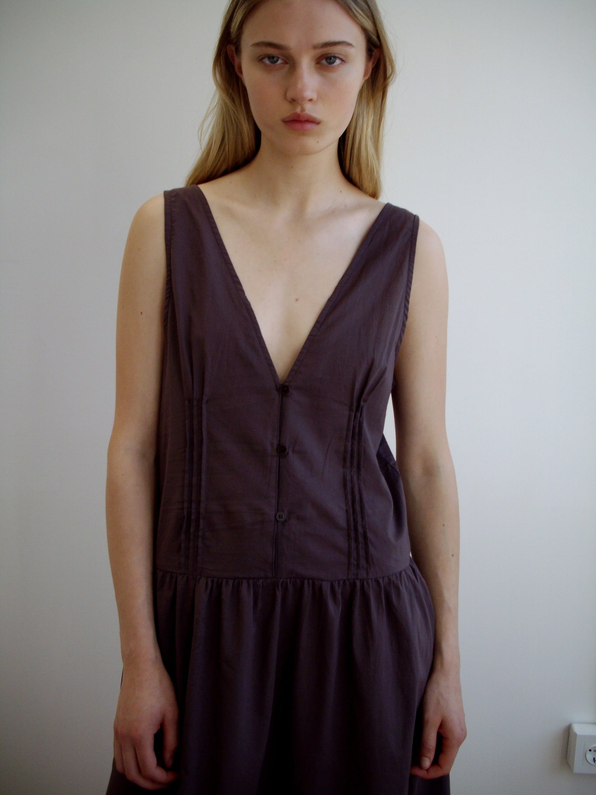 A woman with straight blonde hair wears the Deiji Studios Pintuck Dress in Raisin, a sleeveless deep V-neck style. She stands against a plain white background, gazing at the camera with a neutral expression.