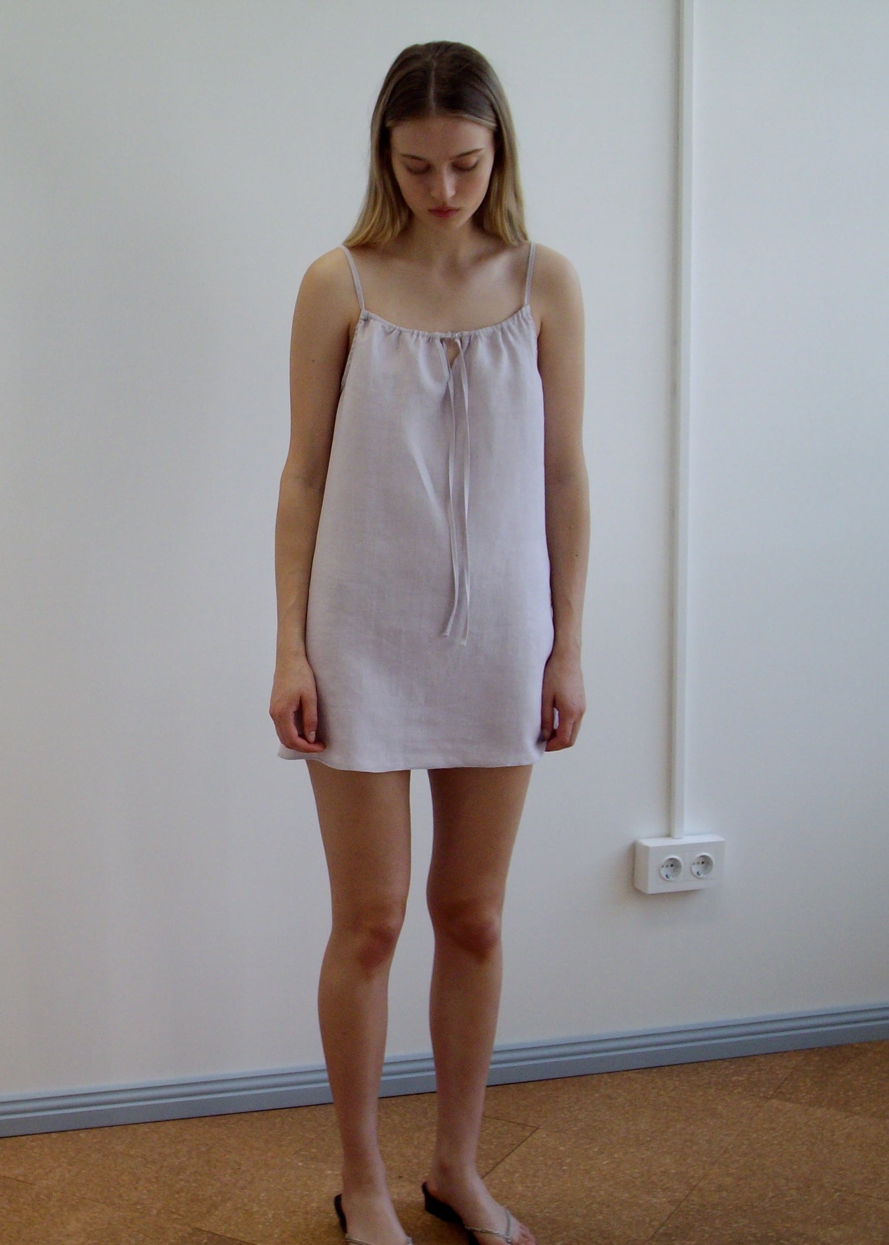 A woman stands indoors against a white wall, wearing the Lettuce Mini Dress - Lilac by Deiji Studios. The sleeveless dress features thin straps, a tie at the neckline, and is crafted from light organic linen. She looks down with a neutral expression.