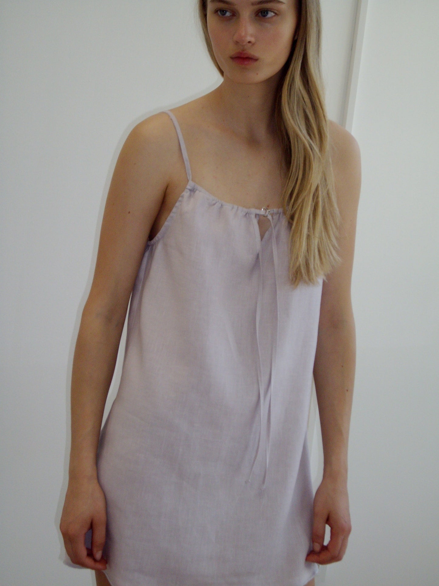 A woman with long blonde hair wears the Lettuce Mini Dress - Lilac by Deiji Studios, an organic linen dress with thin straps and a tie neckline, standing against a plain white background.