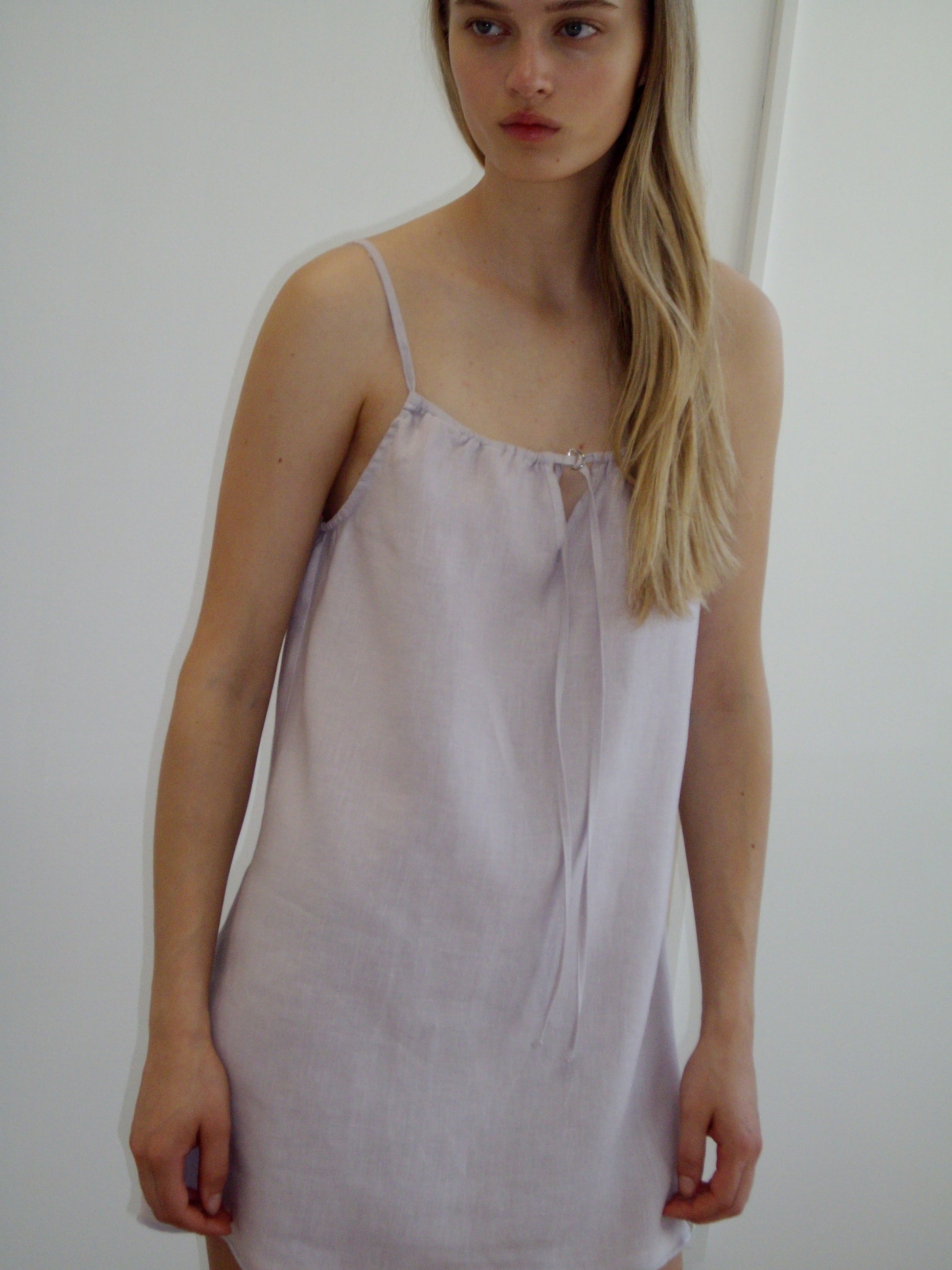 A woman with long blonde hair wears the Lettuce Mini Dress - Lilac by Deiji Studios, an organic linen dress with thin straps and a tie neckline, standing against a plain white background.