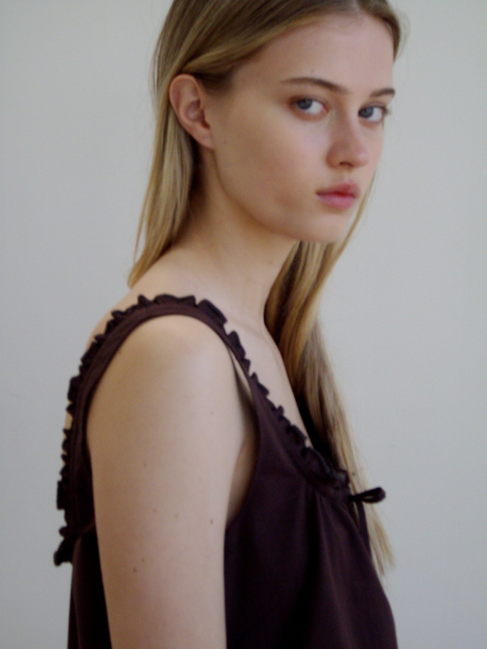 A young woman with straight, long blonde hair wears the Button Frill Mini Dress - Umber by Deiji Studios, standing in profile and looking over her shoulder with a neutral expression against a plain light background.