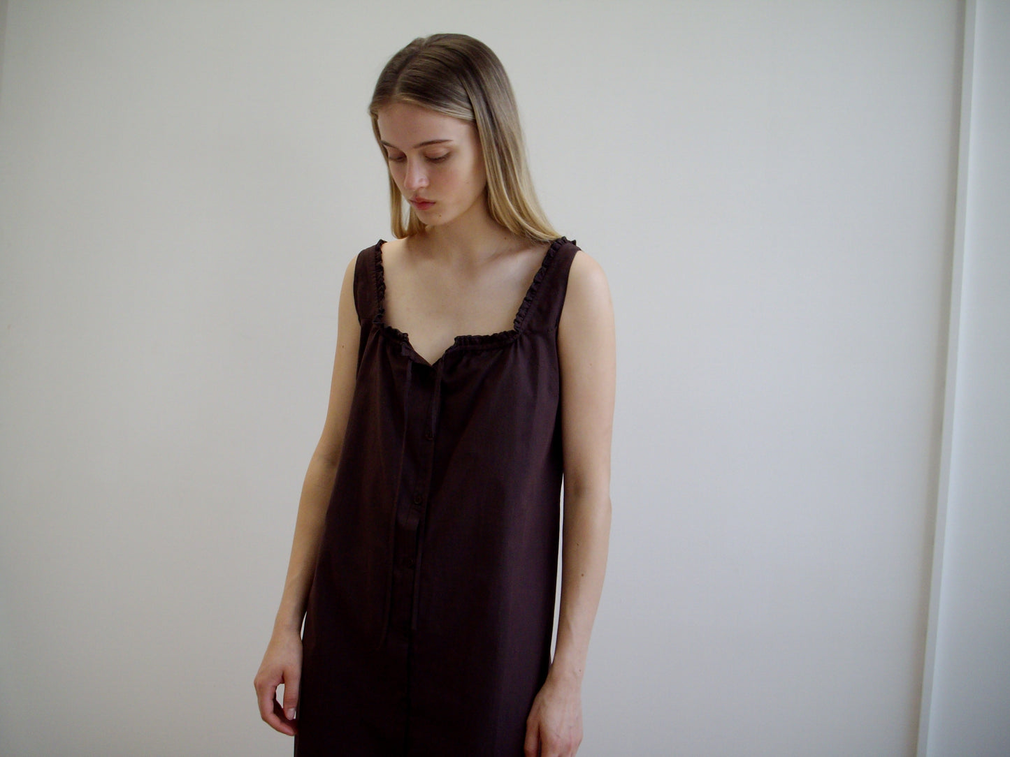 A woman with long, straight, light brown hair stands against a white wall, wearing the Button Sweep Dress - Umber, a sleeveless dark brown organic cotton dress, and gazes downward with a neutral expression.