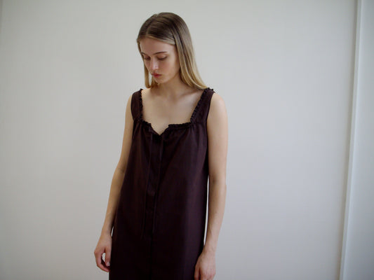 A woman with long, straight, light brown hair stands against a white wall, wearing the Button Sweep Dress - Umber, a sleeveless dark brown organic cotton dress, and gazes downward with a neutral expression.