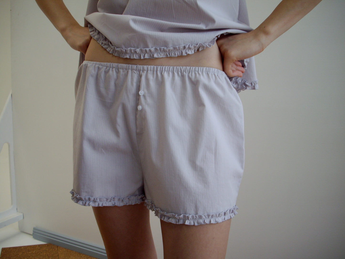 A person stands indoors with hands on hips, wearing the Yoke Set - Lilac Stripe by Deiji Studios, featuring light-colored, loose-fitting pajama shorts and a matching babydoll top with ruffled edges in organic cotton poplin. The background is simple.