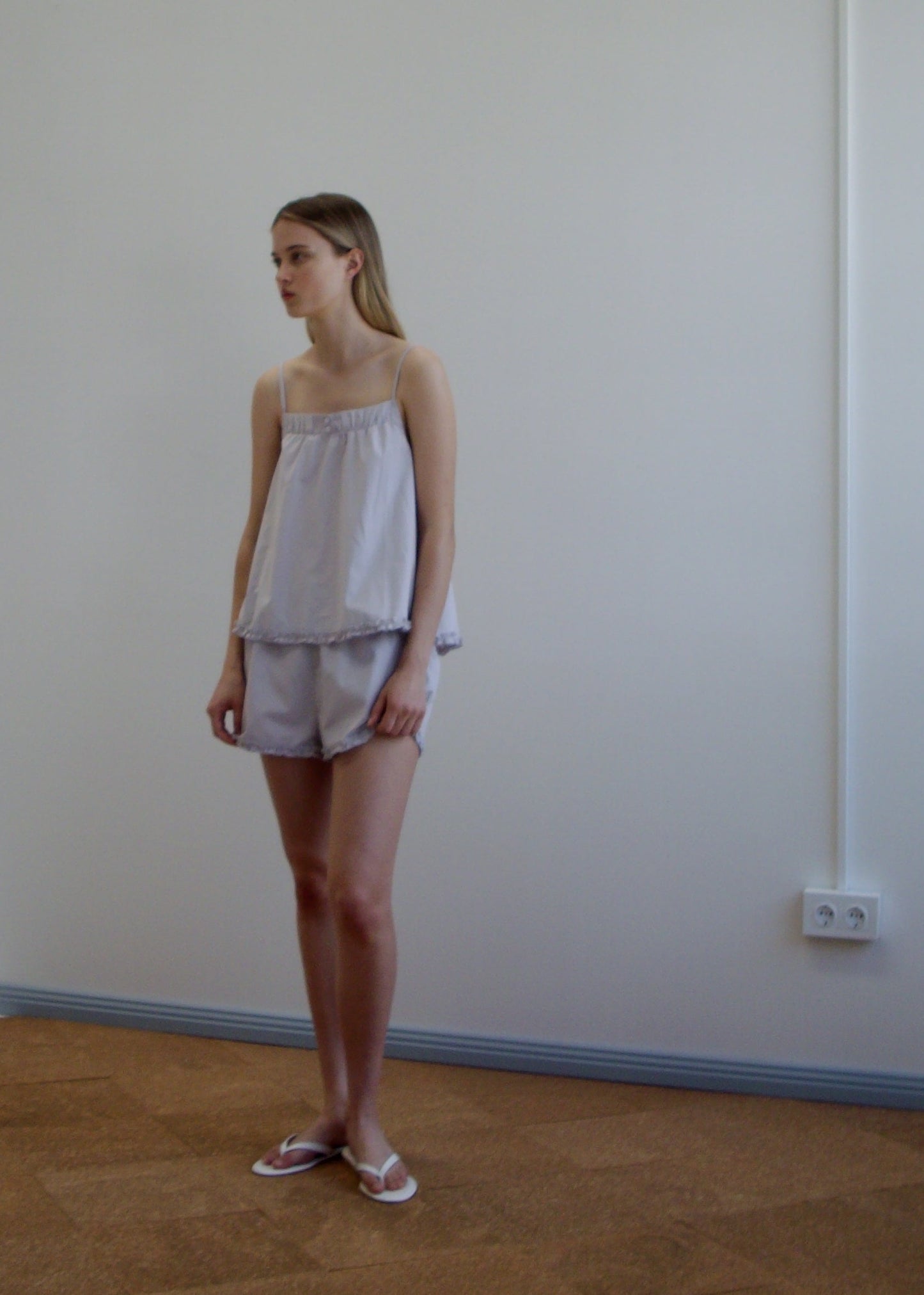 A woman stands indoors against a plain white wall, wearing the Yoke Set - Lilac Stripe by Deiji Studios with slippers. She has straight shoulder-length hair and looks to the side.