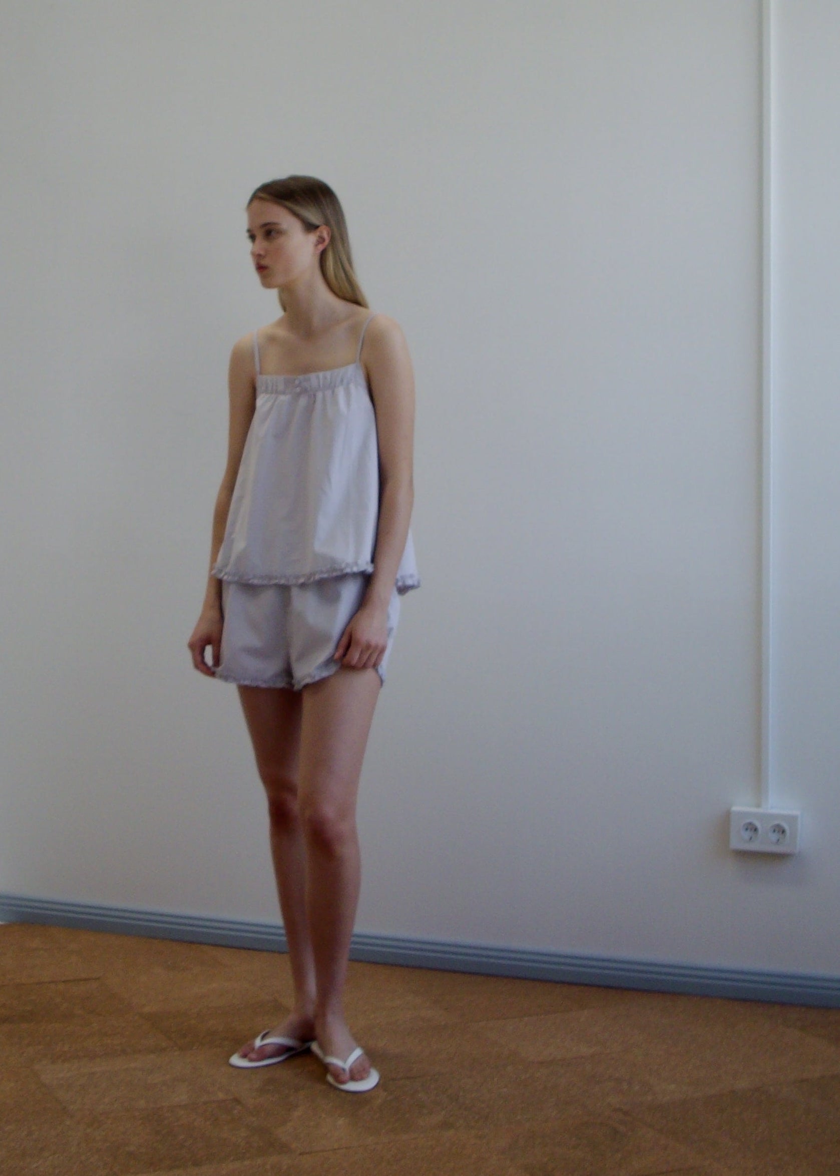 A woman stands indoors against a plain white wall, wearing the Yoke Set - Lilac Stripe by Deiji Studios with slippers. She has straight shoulder-length hair and looks to the side.