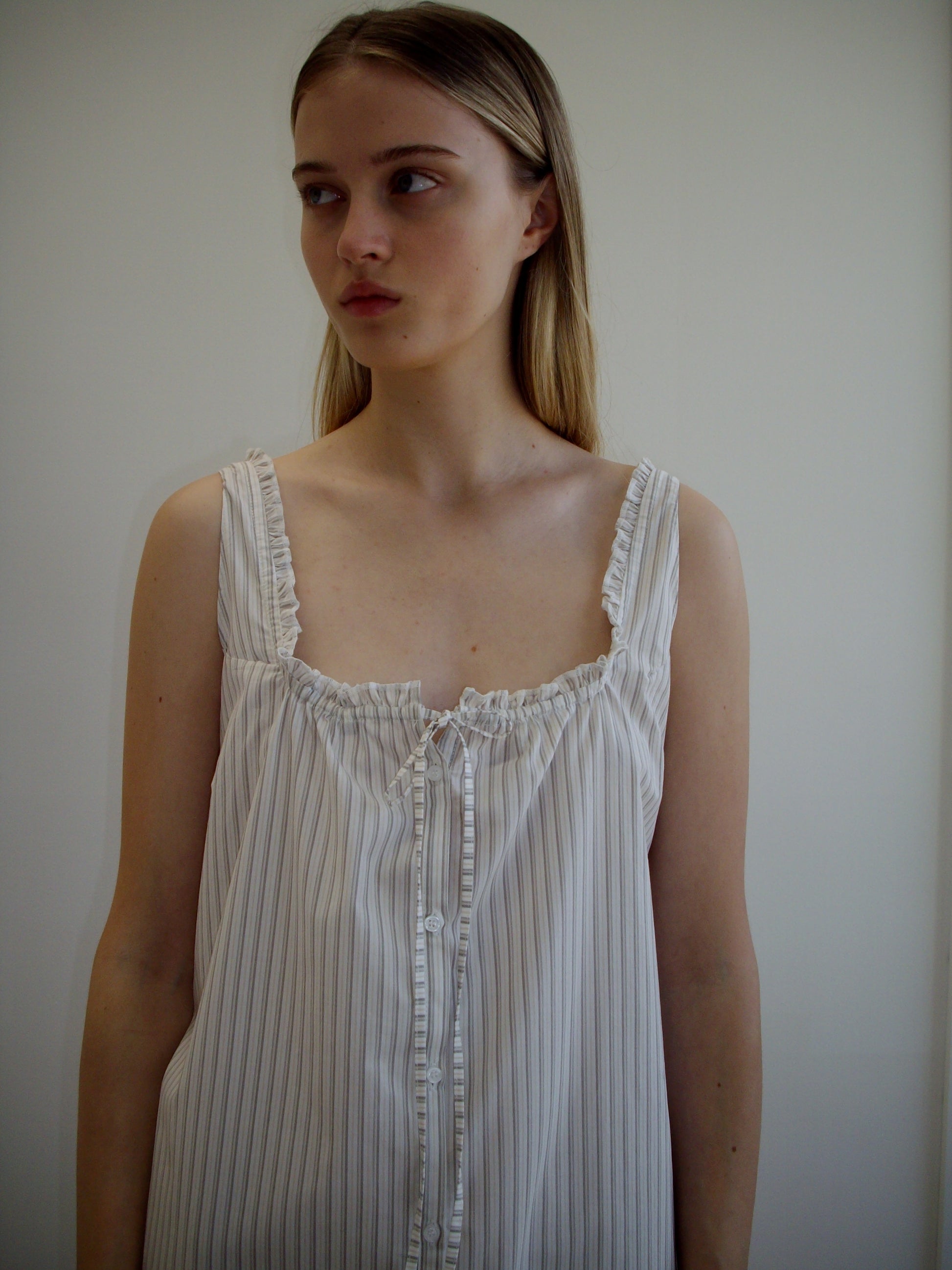 A young woman with long, straight hair wears the Button Sweep Dress - Gull Stripe, a loose, sleeveless white and gray striped organic cotton dress with ruffles, standing against a plain light background and looking slightly to the side.