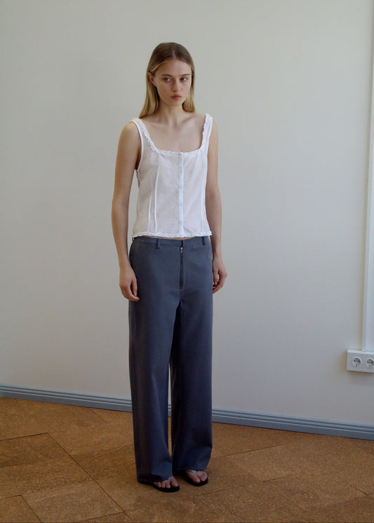 A woman stands indoors against a plain wall, wearing Deiji Studios’ white sleeveless top and the Zip Pant - Charcoal in organic cotton twill. She has long, straight hair and a neutral expression; the brown floor has a visible power outlet.