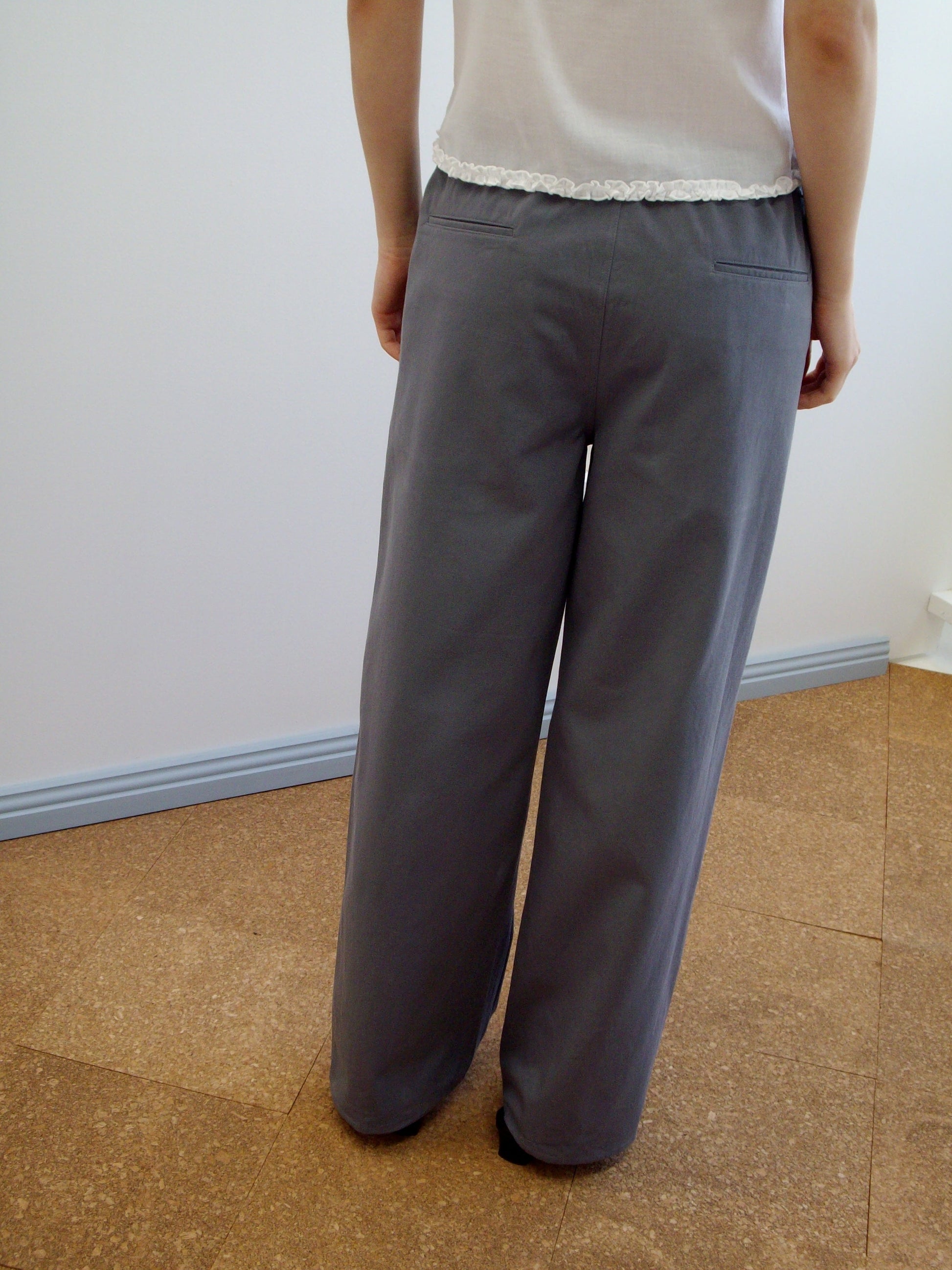 A person stands indoors on a cork floor, facing away from the camera, wearing loose-fitting Deiji Studios Zip Pant - Charcoal and a white short-sleeve top with a ruffled hem.