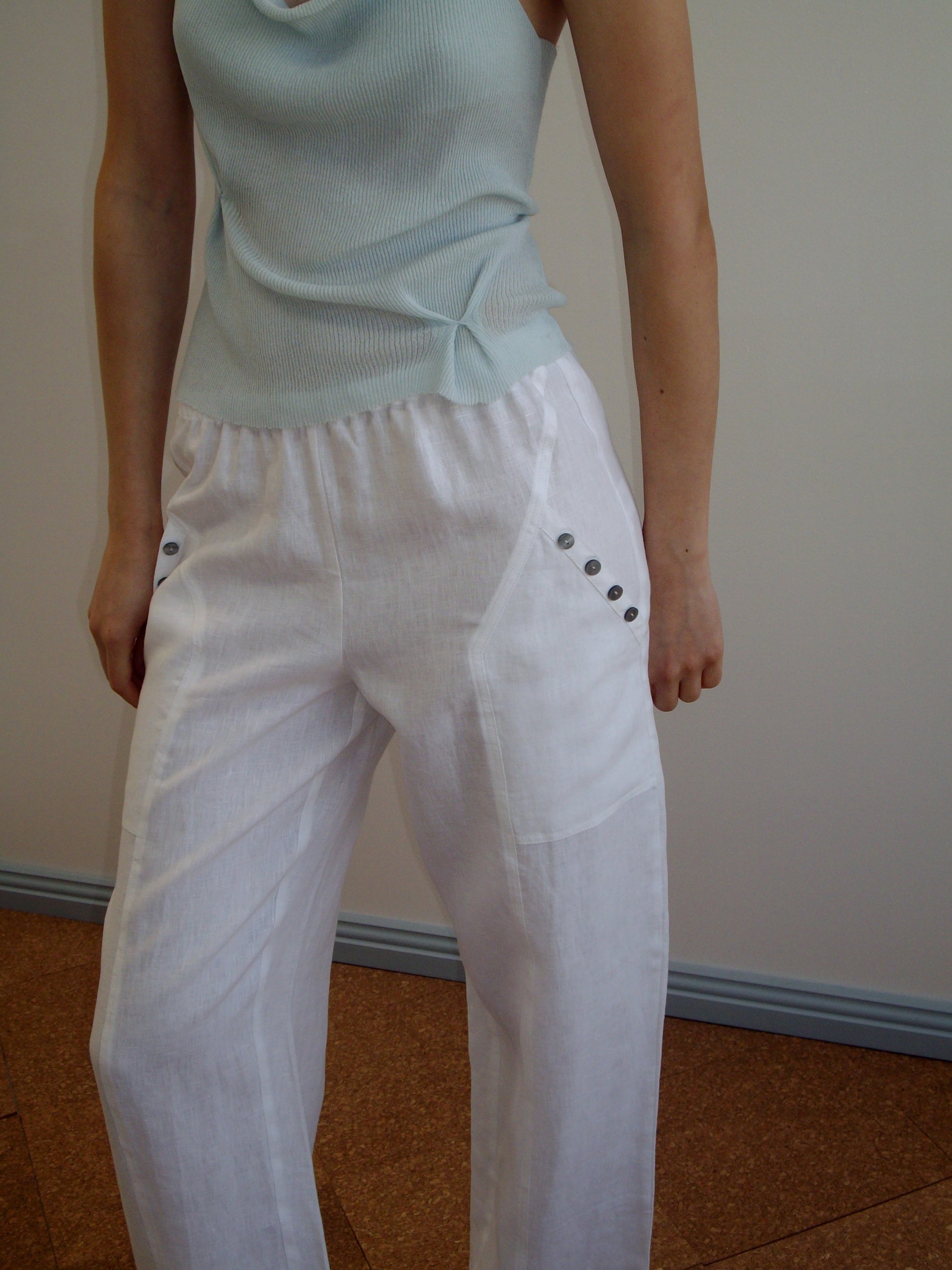 A person wears a sleeveless light blue top and the Angle Pocket Pant - White by Deiji Studios, featuring relaxed organic linen and button details at the pockets, standing against a plain wall with their upper body and legs visible.