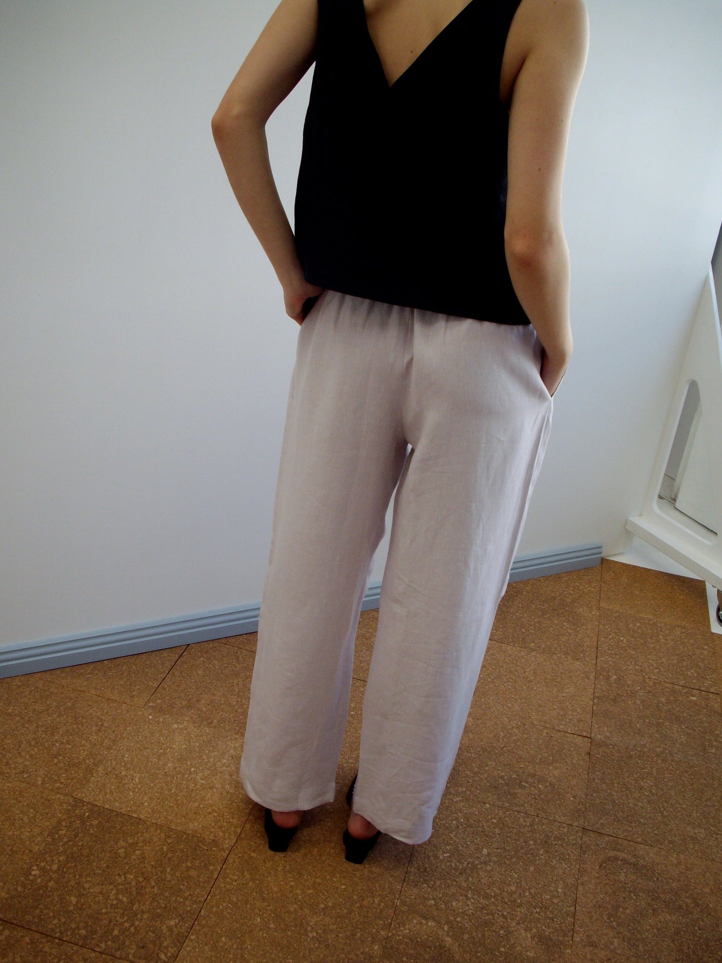 A person stands indoors with their back to the camera, wearing a sleeveless black top and Linen Ease Trouser in lilac, hands tucked in the pockets.