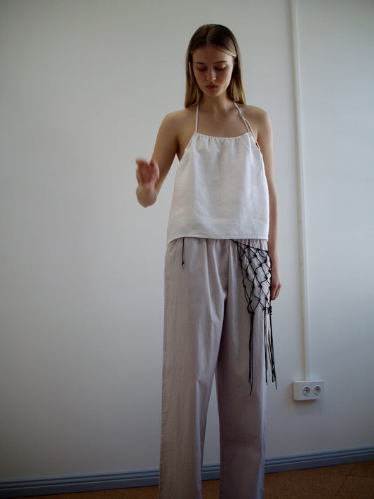 A woman stands indoors against a plain white wall, wearing a loose organic cotton halter top by Deiji Studios and Ease Trouser - Lilac Stripe with a black netted accessory on the right. She looks down, one hand slightly raised.
