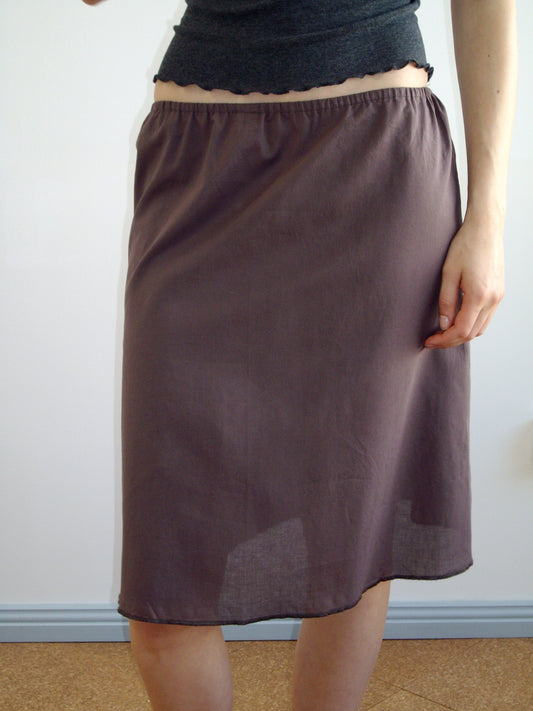 A person wearing a dark gray top and the knee-length, loose-fitting Simple Skirt - Raisin stands against a plain light-colored wall.