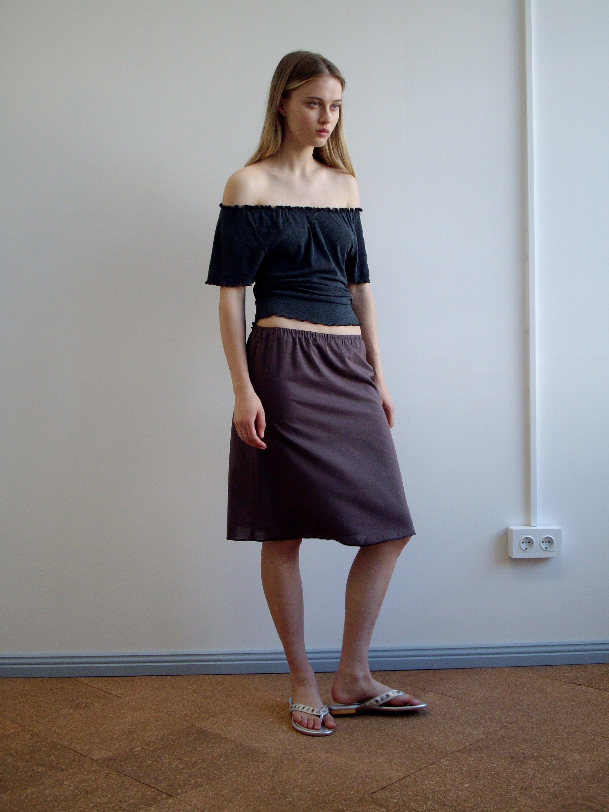 A woman with long hair and a neutral expression stands indoors against a plain white wall, wearing an off-the-shoulder black top, the Simple Skirt - Raisin, and white sandals.