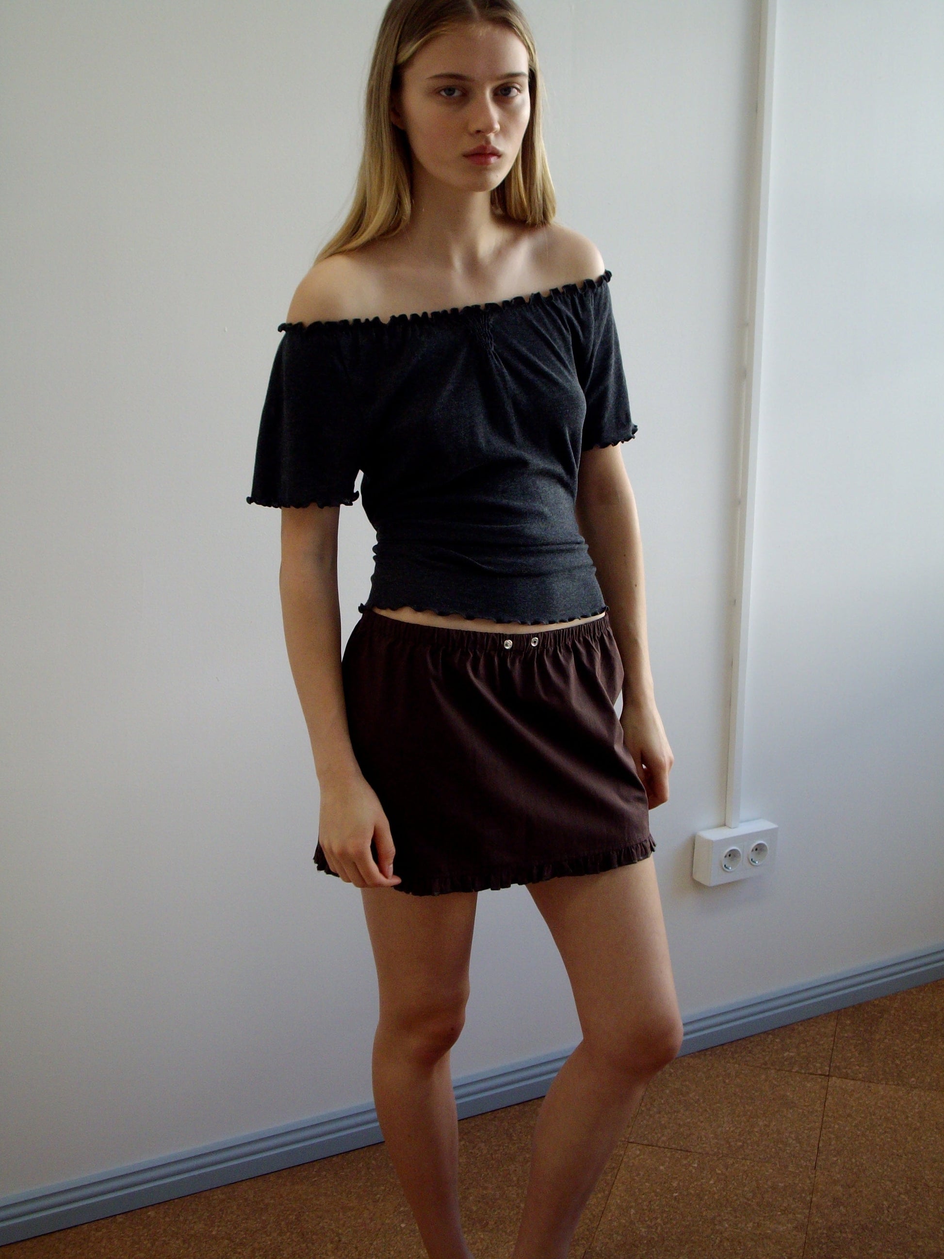 A young woman with straight blonde hair wears an off-the-shoulder dark top and the Deiji Studios Flounce Mini - Umber skirt. She stands indoors against a plain white wall with a power outlet visible behind her.