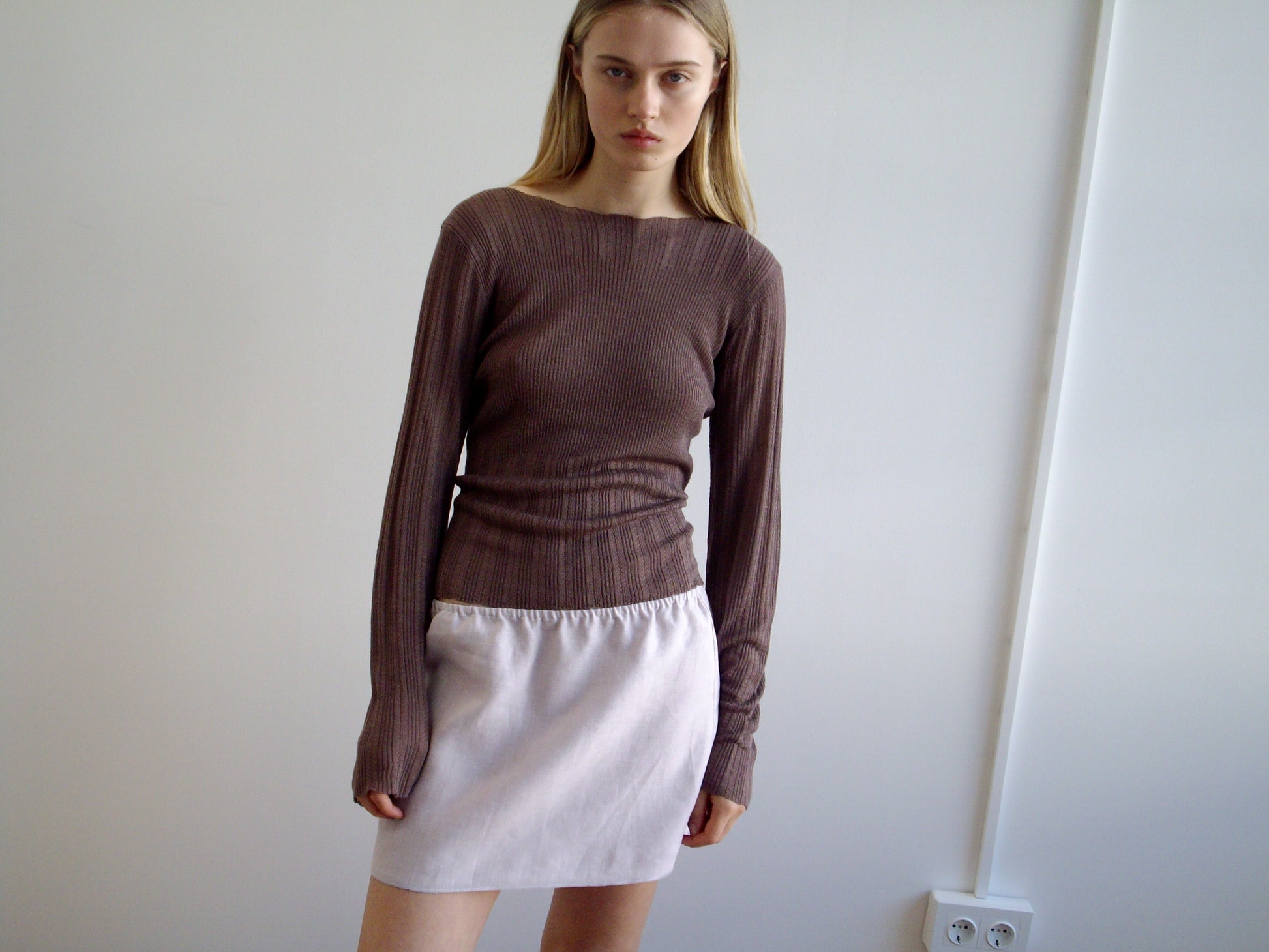 A woman with long blonde hair stands indoors, wearing a fitted brown long-sleeve top and the Babylock Skirt - Lilac. She poses before a plain white wall with an electrical outlet visible.