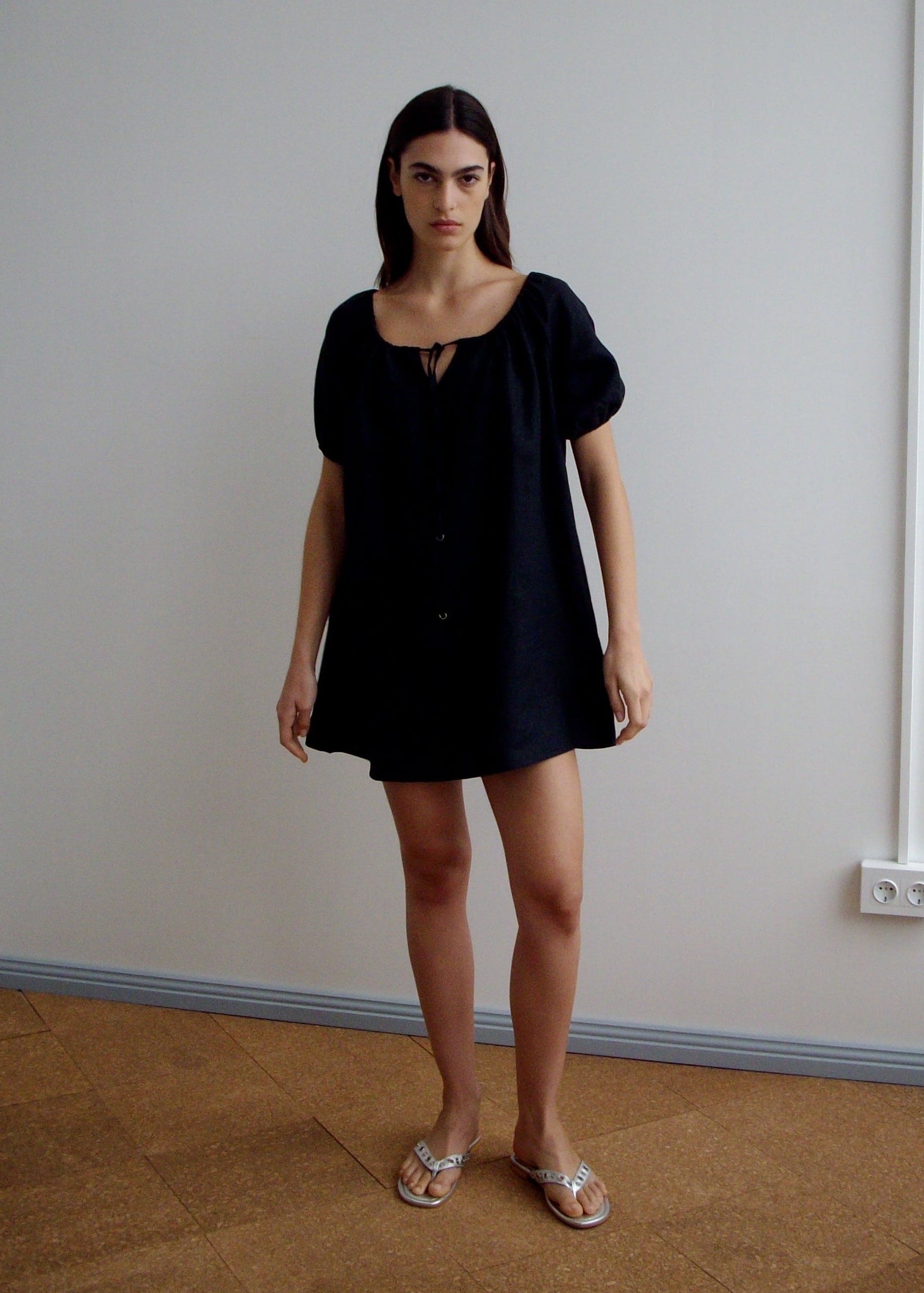 A woman with long dark hair stands indoors against a plain wall, wearing Deiji Studios' loose-fitting Smock Dress - Black with short sleeves, a neckline tie, and flat metallic sandals.