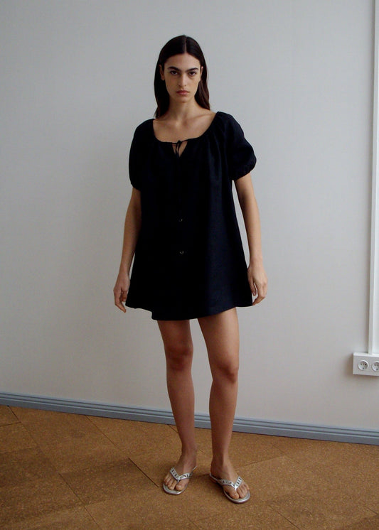 A woman with long dark hair stands indoors against a plain wall, wearing Deiji Studios' loose-fitting Smock Dress - Black with short sleeves, a neckline tie, and flat metallic sandals.
