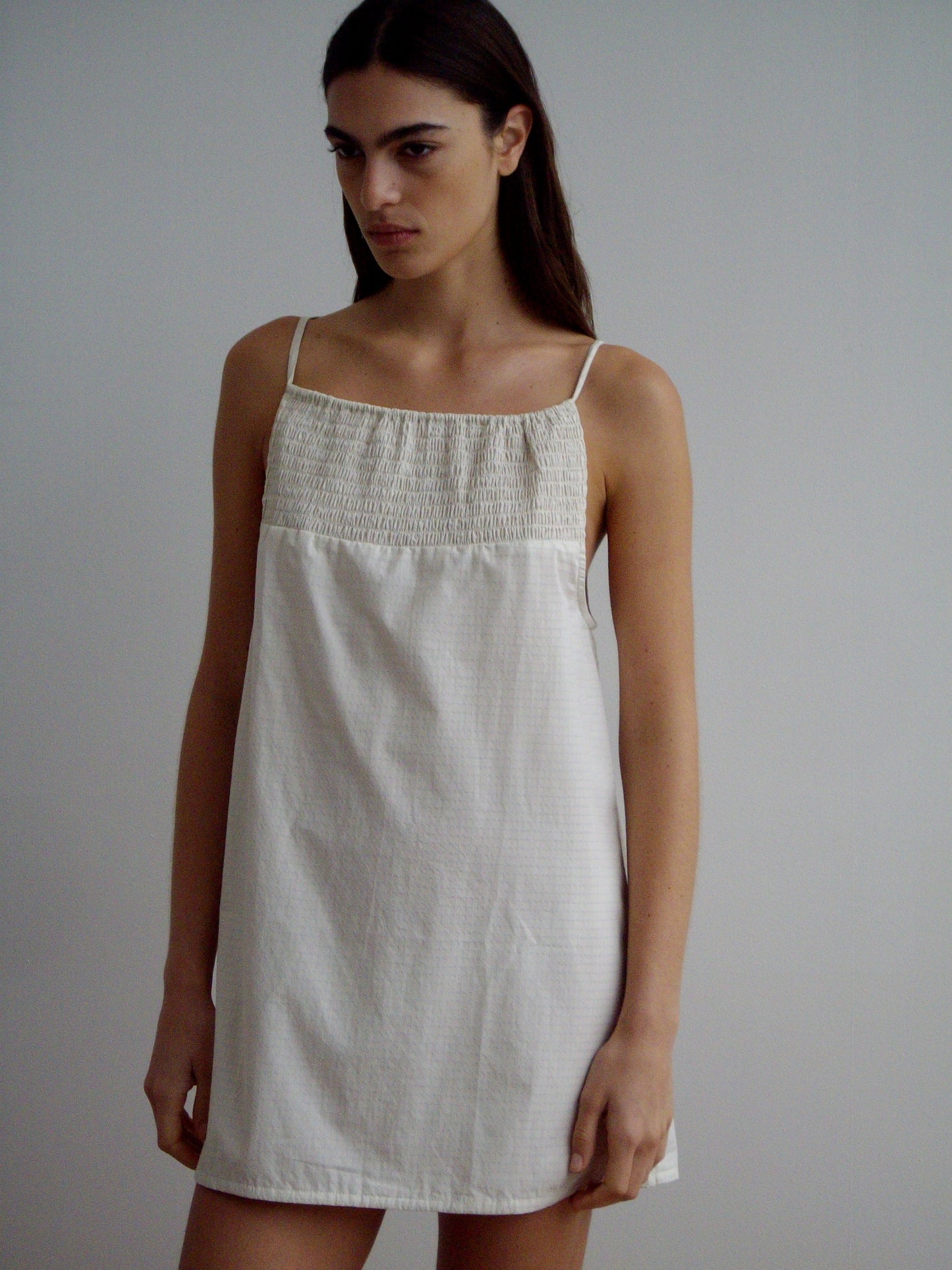 A woman with long, straight brown hair wears the Shirring Mini Dress - Bone Check by Deiji Studios—a sleeveless, white shirred mini dress with thin straps and a gathered neckline—standing against a plain light background.