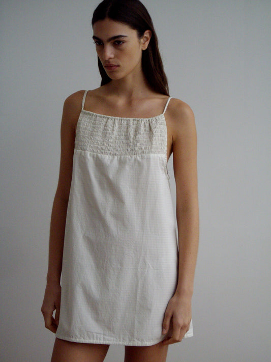 A woman with long, straight brown hair wears the Shirring Mini Dress - Bone Check by Deiji Studios—a sleeveless, white shirred mini dress with thin straps and a gathered neckline—standing against a plain light background.
