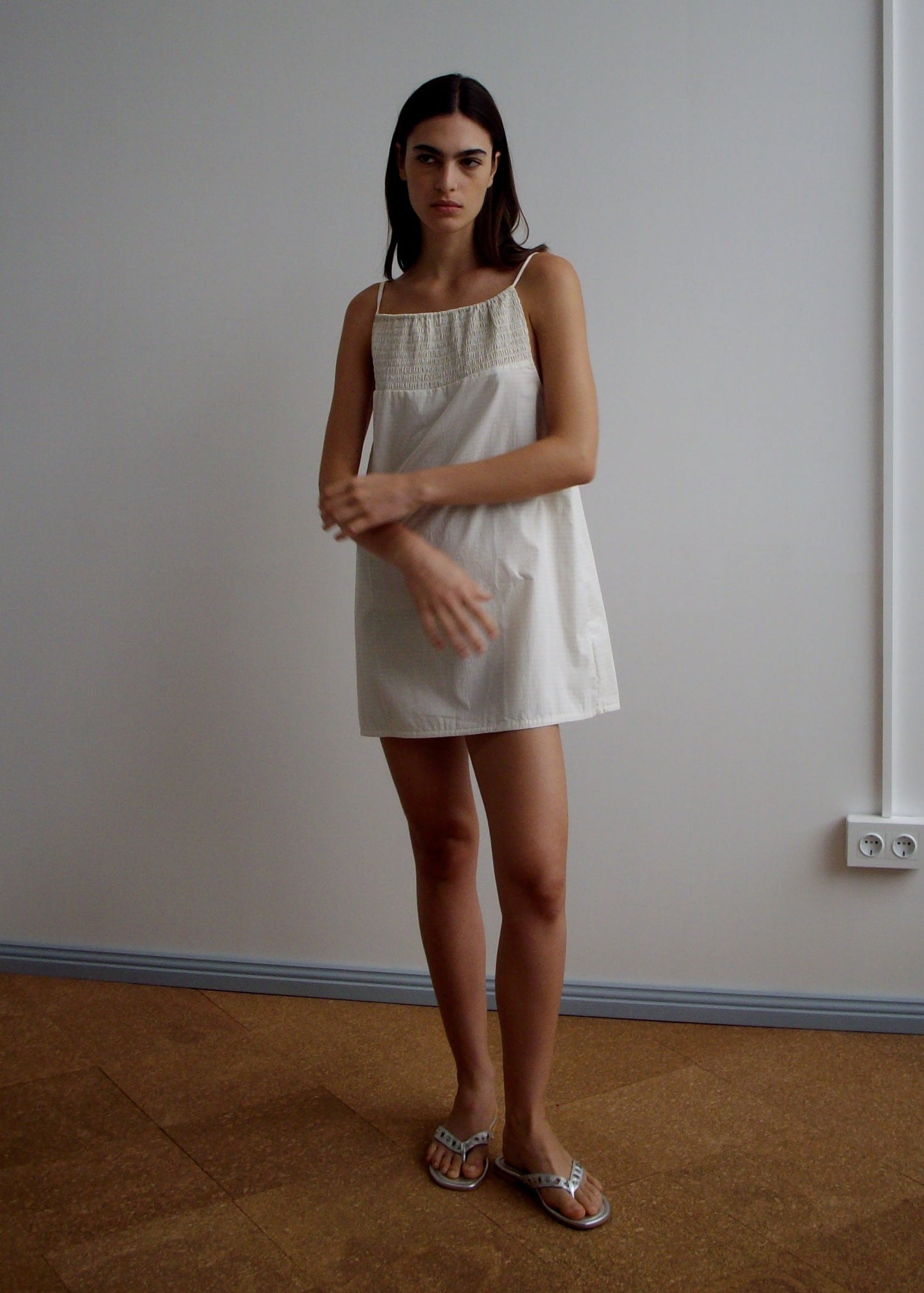 A woman with long dark hair stands indoors against a light wall, wearing the Shirring Mini Dress - Bone Check and sandals. She has a neutral expression and her hands are raised near her chest.