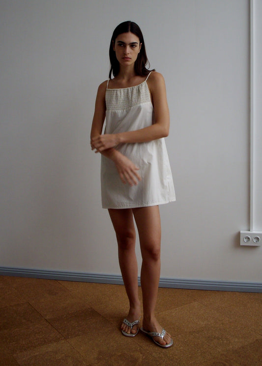 A woman with long dark hair stands indoors against a light wall, wearing the Shirring Mini Dress - Bone Check and sandals. She has a neutral expression and her hands are raised near her chest.