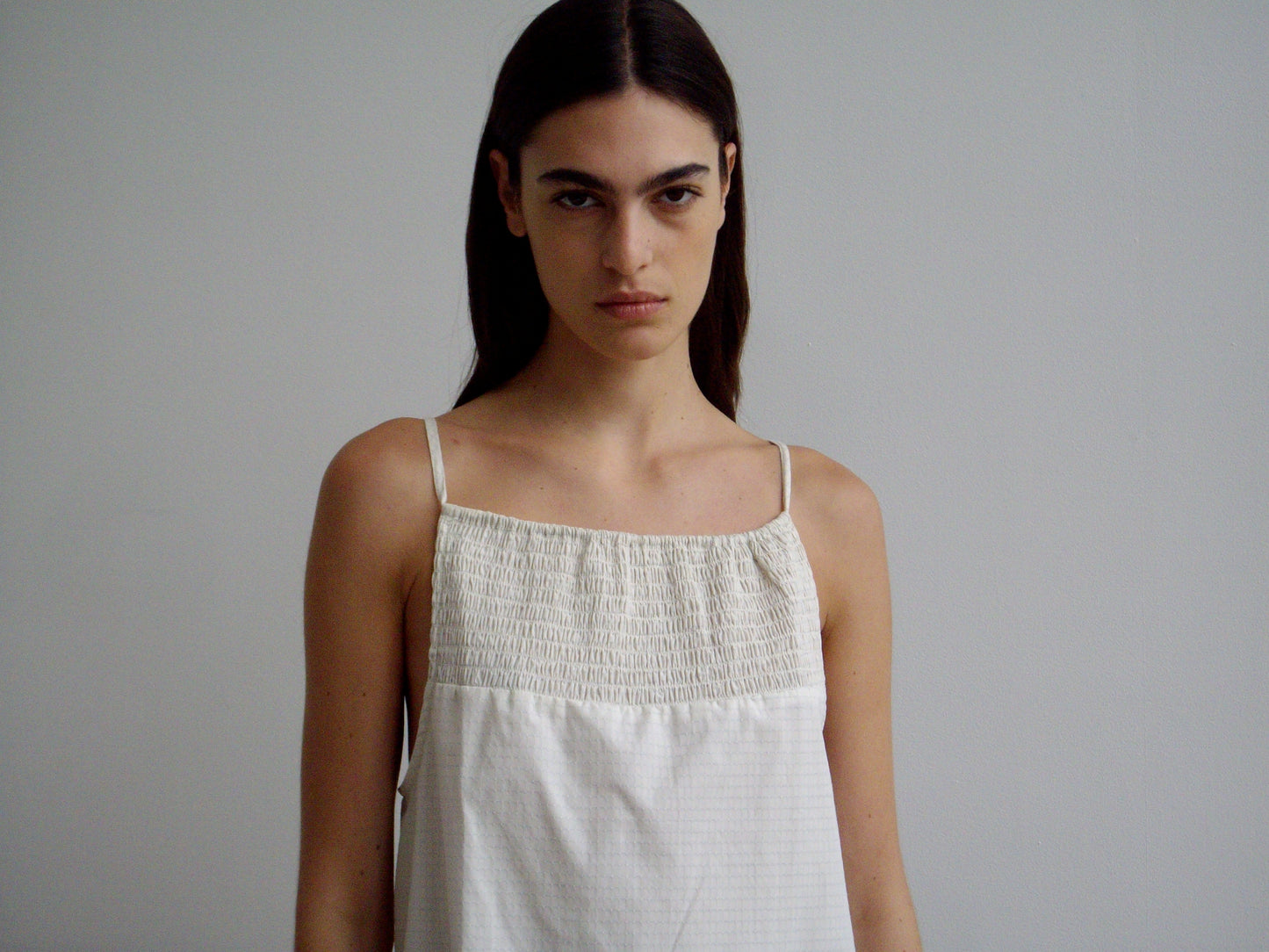A person with long dark hair wears the Shirring Mini Dress - Bone Check, featuring thin straps and a smocked top, standing against a plain light gray background and looking directly at the camera with a neutral expression.