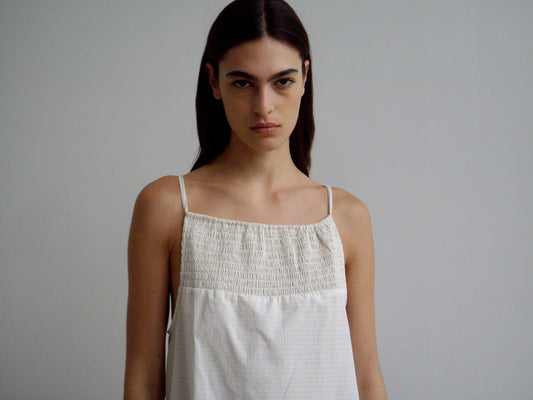 A person with long dark hair wears the Shirring Mini Dress - Bone Check, featuring thin straps and a smocked top, standing against a plain light gray background and looking directly at the camera with a neutral expression.