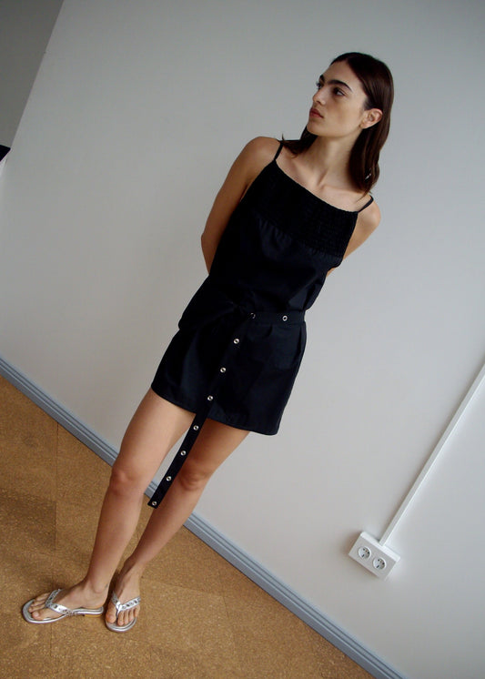 A woman stands indoors against a white wall, wearing the Shirring Mini Dress - Black by Deiji Studios. The sleeveless organic cotton dress features button details and is paired with sandals. She looks to the side with arms behind her back.