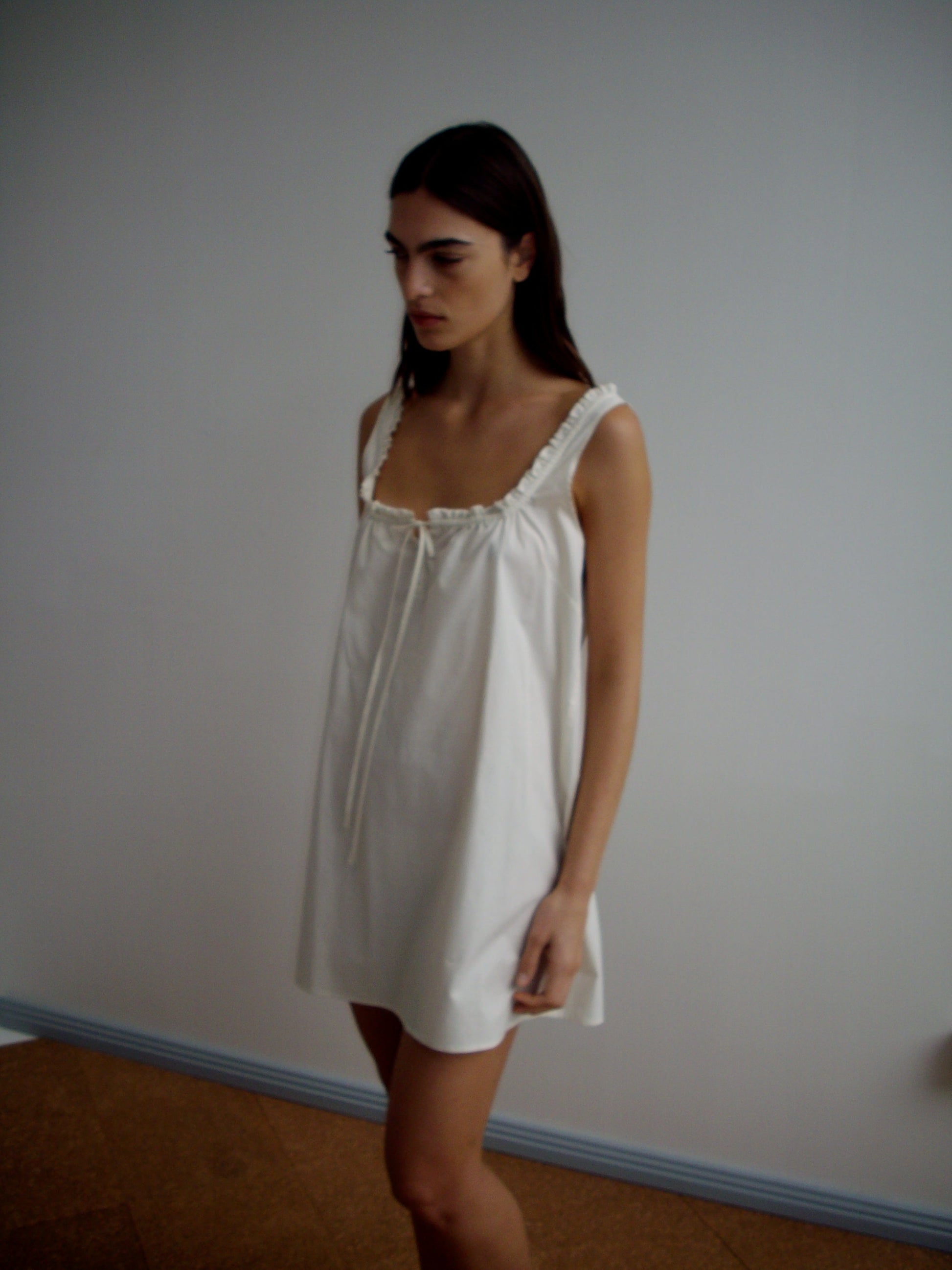 A woman with long dark hair stands indoors against a plain light wall, wearing the Button Frill Mini Dress - Stone Stripe by Deiji Studios, a loose organic cotton piece with ruffled straps.