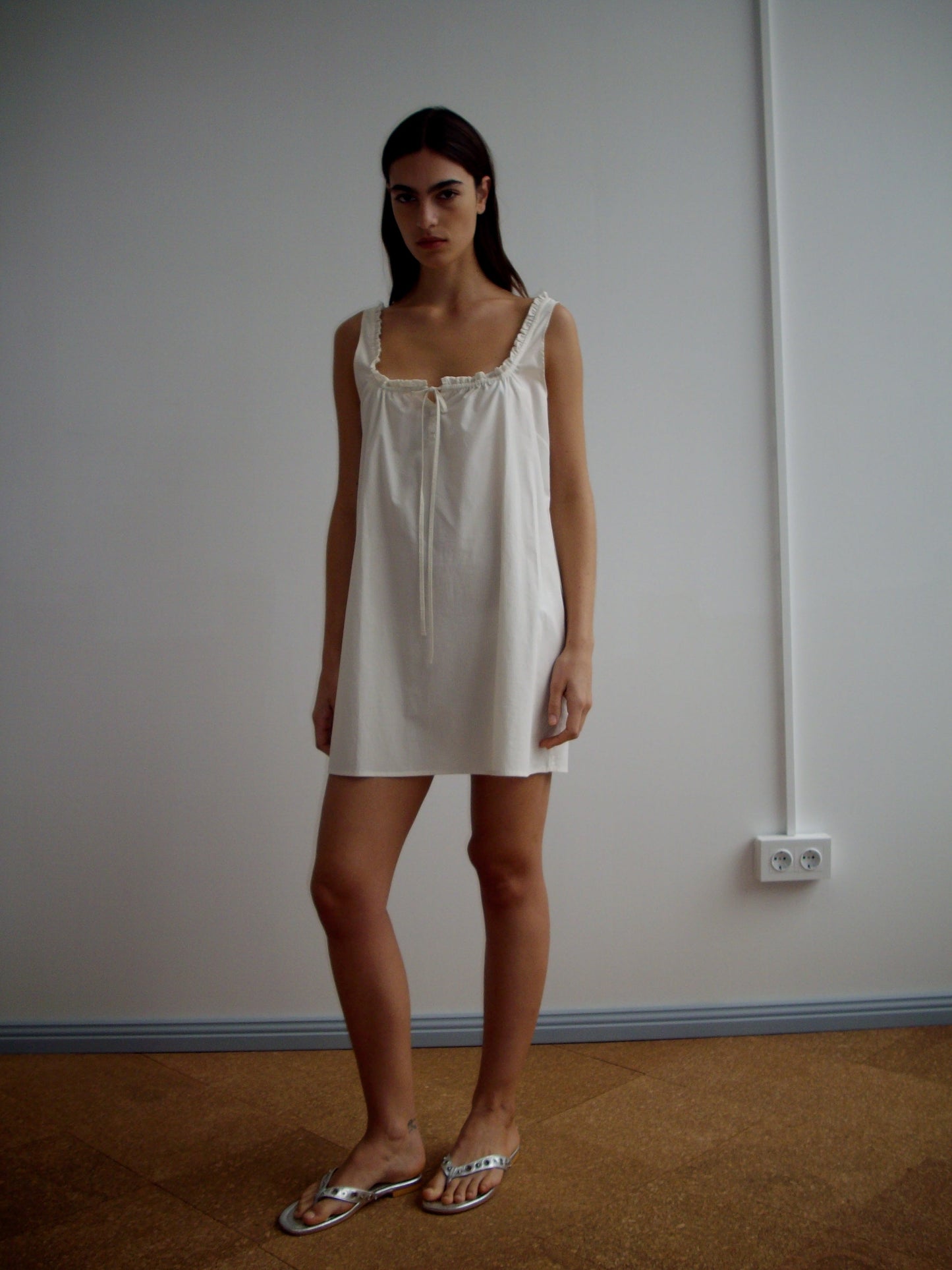 A woman with straight dark hair and a neutral expression stands indoors against a plain white wall, wearing the Button Frill Mini Dress - Stone Stripe and silver sandals, with her arms relaxed by her sides.