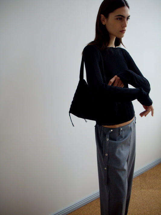 A person with long dark hair stands against a white wall, wearing a black long-sleeve top and grey blue stripe Wool Belt Pant with visible buttons. They carry a black shoulder bag, arms crossed, and have a neutral expression.