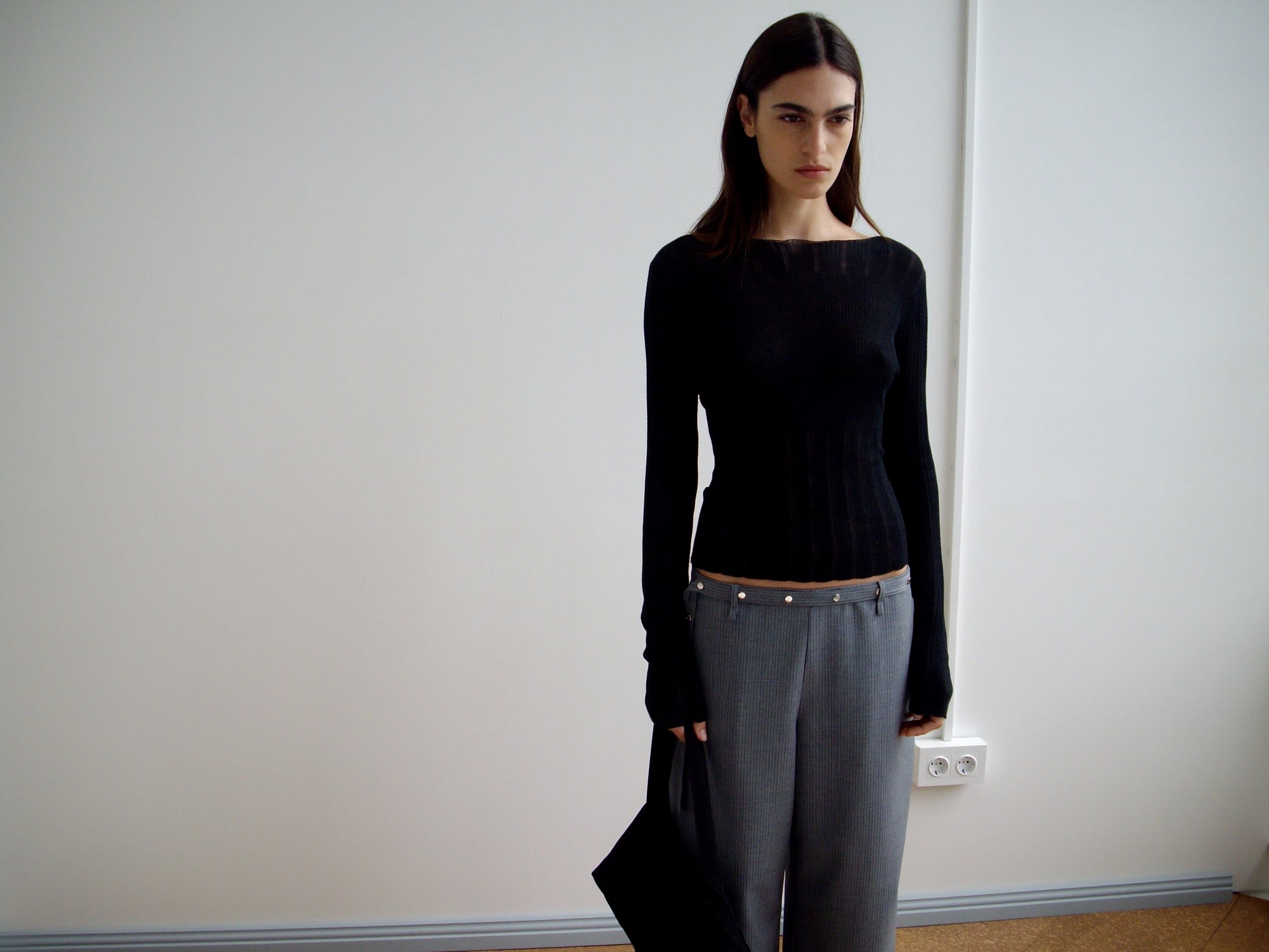 A woman with straight dark hair wears a fitted black long-sleeve top and Wool Belt Pant in Grey Blue Stripe, standing indoors against a plain white wall while holding a black bag at her side.