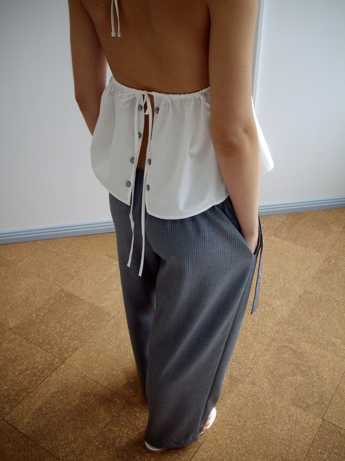 A person stands with their back turned, wearing Deiji Studios' Button Halter - White, an organic cotton backless halter top, paired with loose-fitting gray pinstriped pants and hands casually in their pockets.