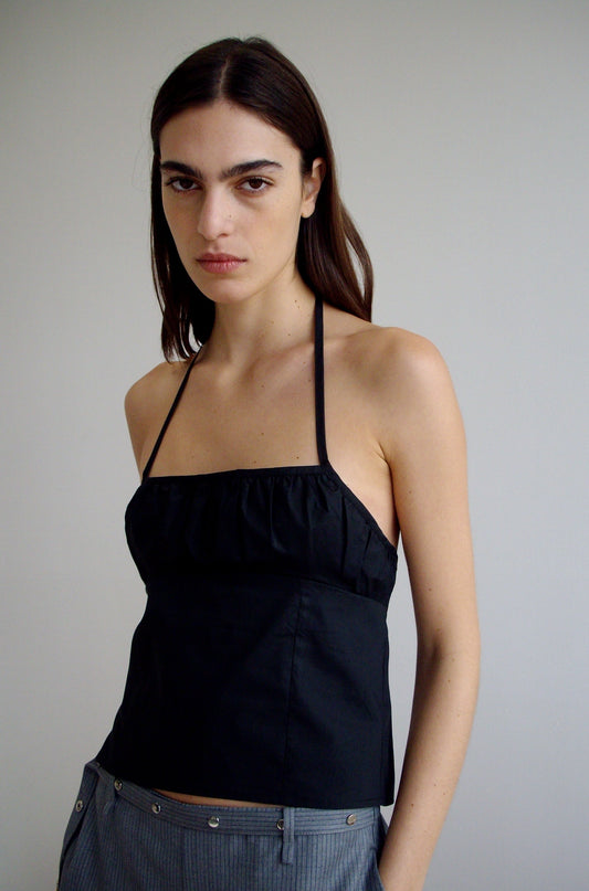 A woman with straight brown hair and strong eyebrows stands against a plain light wall, wearing Deiji Studios' Strap Top - Black and gray organic cotton pants, with her hands in her pockets.