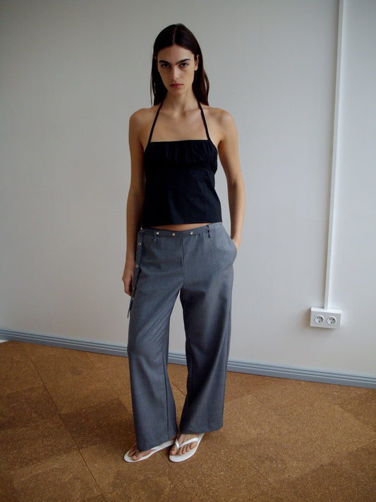 A woman stands indoors against a plain wall, wearing a black halter top and Wool Belt Pant - Grey Blue Stripe with white sandals. She has long dark hair, a serious expression, and her hands in the pockets of her pants.