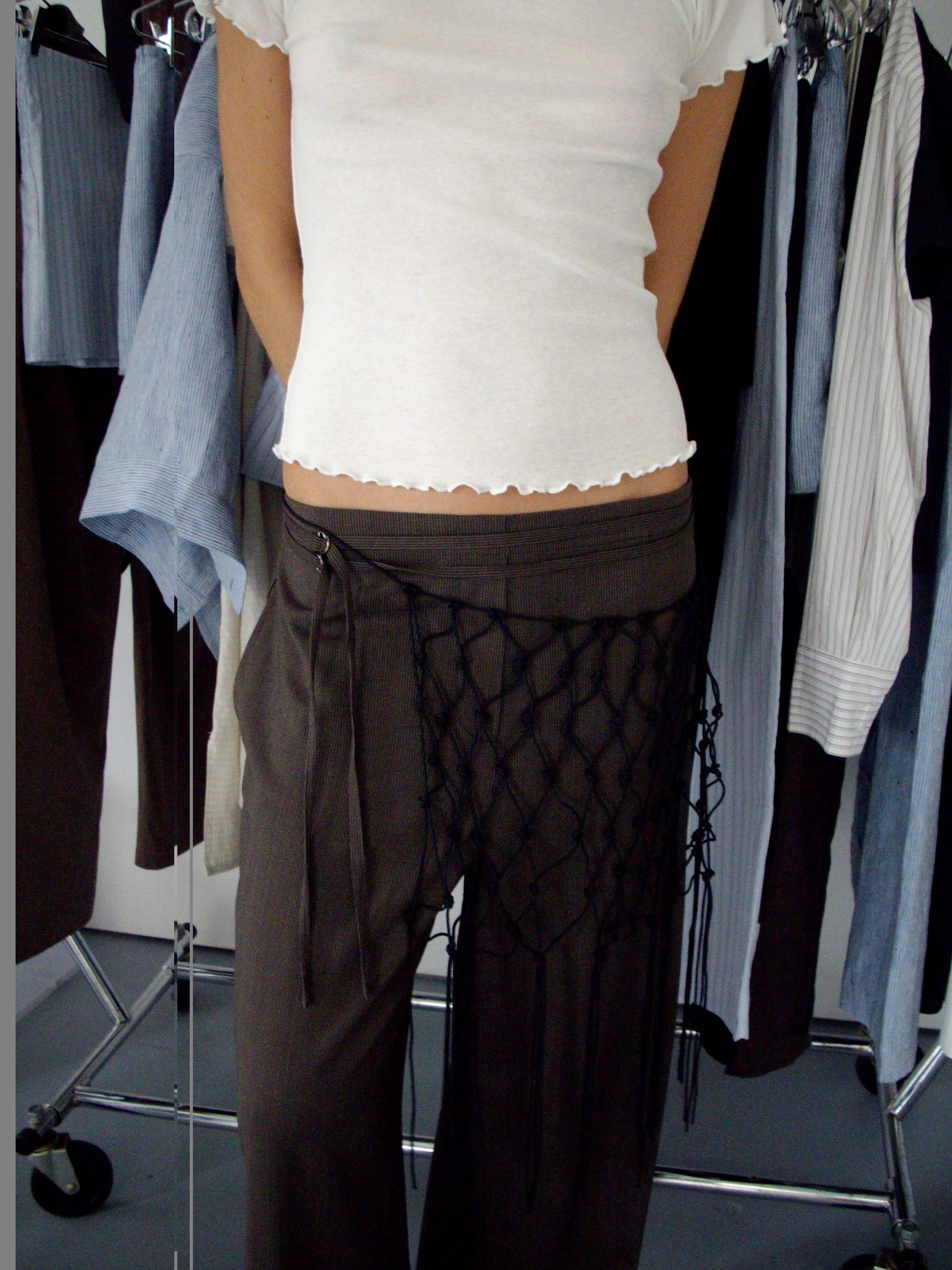 A person wearing a white short-sleeved top and the Double Belted Pant - Brown Stripe, accessorized with a black netted piece, stands in front of a rack with various hanging garments.