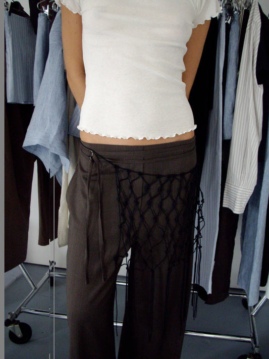 A person wearing a white short-sleeved top and the Double Belted Pant - Brown Stripe, accessorized with a black netted piece, stands in front of a rack with various hanging garments.