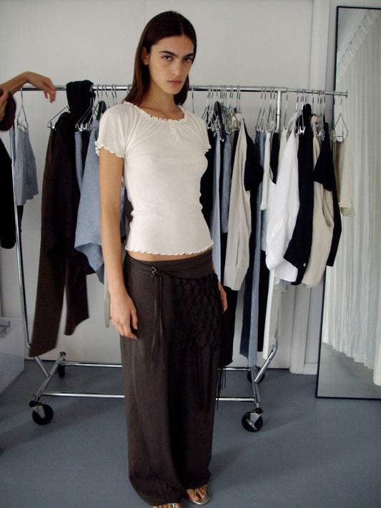 A woman stands indoors before a clothing rack, wearing the Double Belted Pant – Brown Stripe and a white fitted top, accessorized with a black netted piece. She looks at the camera with a neutral expression; clothes hang behind her.