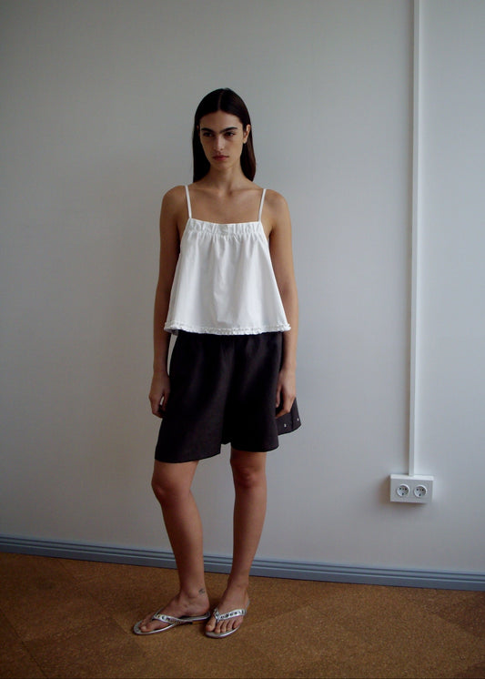 A woman with long dark hair stands indoors against a plain wall, wearing a white sleeveless top and the Press Stud Short - Bark by Deiji Studios. She has sandals on, a neutral expression, and an electrical outlet is visible beside her.