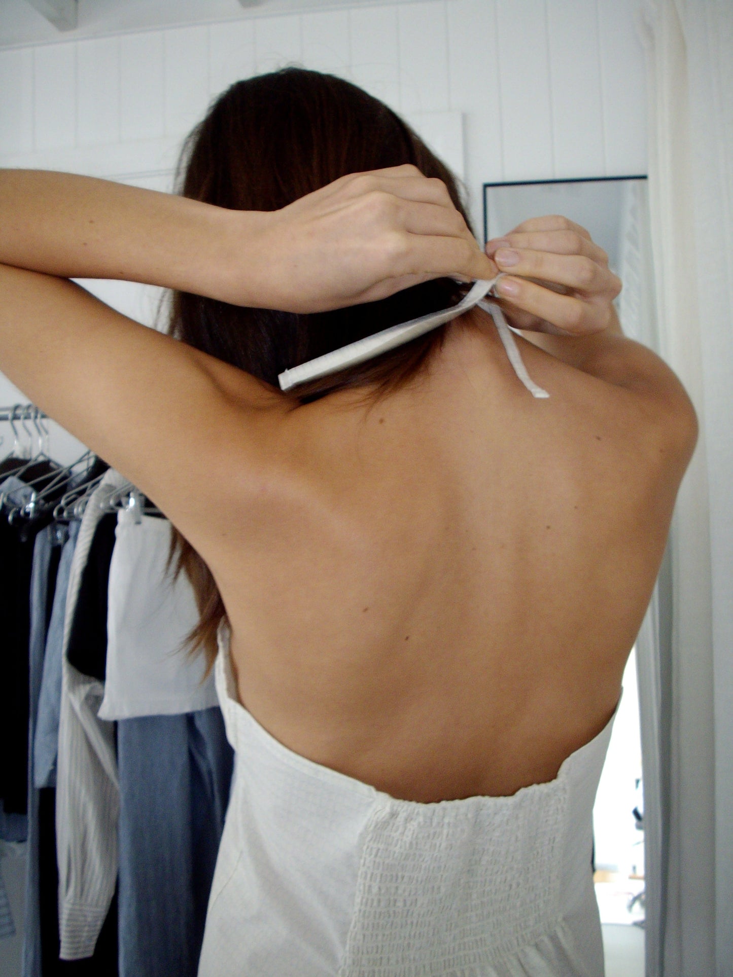 A person with long brown hair ties the ribbon of the Strap Top - Bone Check behind their neck, standing in a room with hanging clothes and a mirror in the background.