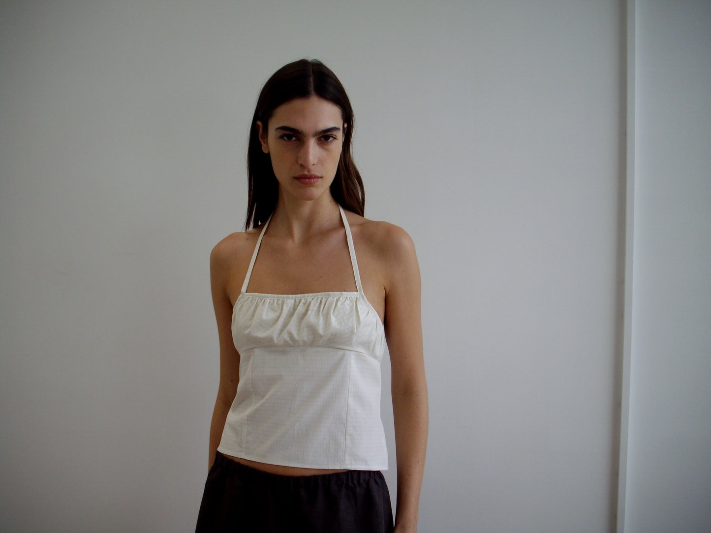 A woman with long brown hair, wearing the Strap Top - Bone Check and dark pants, stands against a plain white wall, looking directly at the camera with a neutral expression.