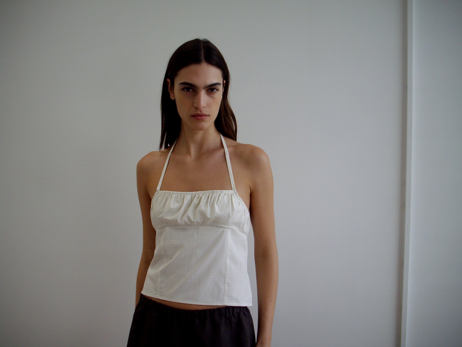 A woman with long brown hair, wearing the Strap Top - Bone Check and dark pants, stands against a plain white wall, looking directly at the camera with a neutral expression.