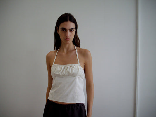 A woman with long brown hair, wearing the Strap Top - Bone Check and dark pants, stands against a plain white wall, looking directly at the camera with a neutral expression.