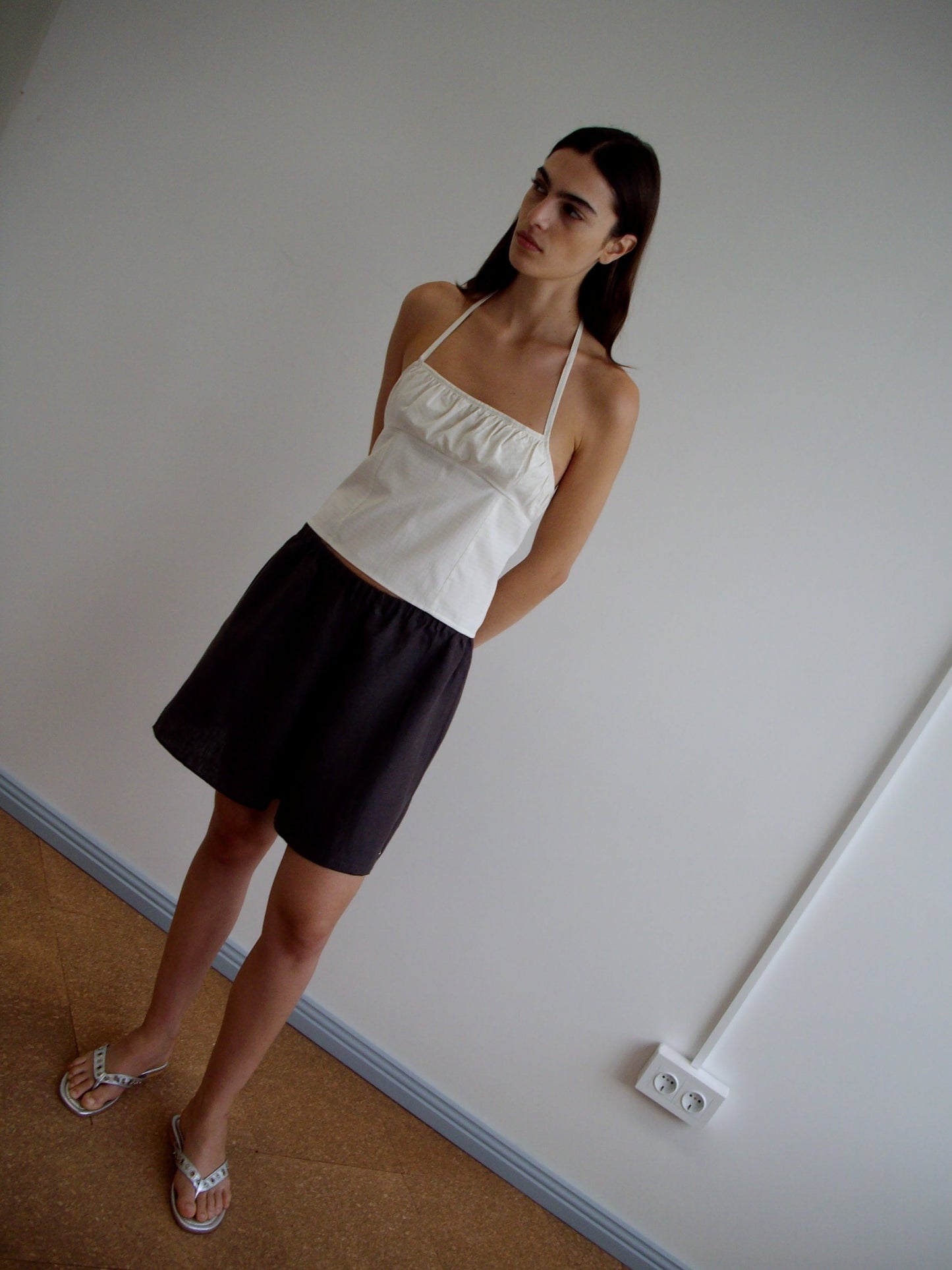A woman with long dark hair stands indoors against a plain white wall, wearing the Strap Top - Bone Check, dark shorts, and flip-flops, with her hands behind her back.