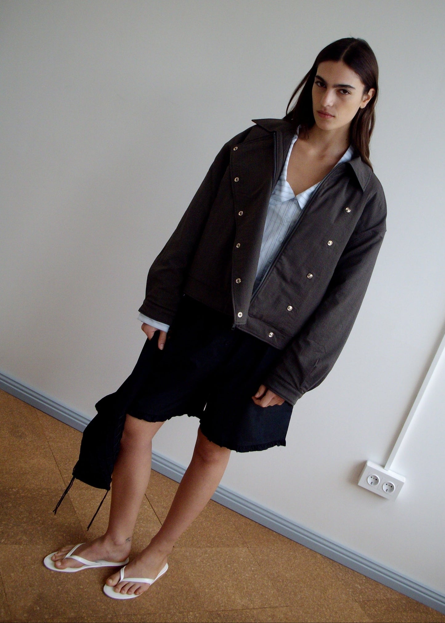 A woman stands indoors against a plain wall, wearing a dark oversized jacket, a light blue shirt, and Deiji Studios’ Flounce Short in black—one side features an asymmetrical, longer flounce hem. She finishes the look with white flip-flops.