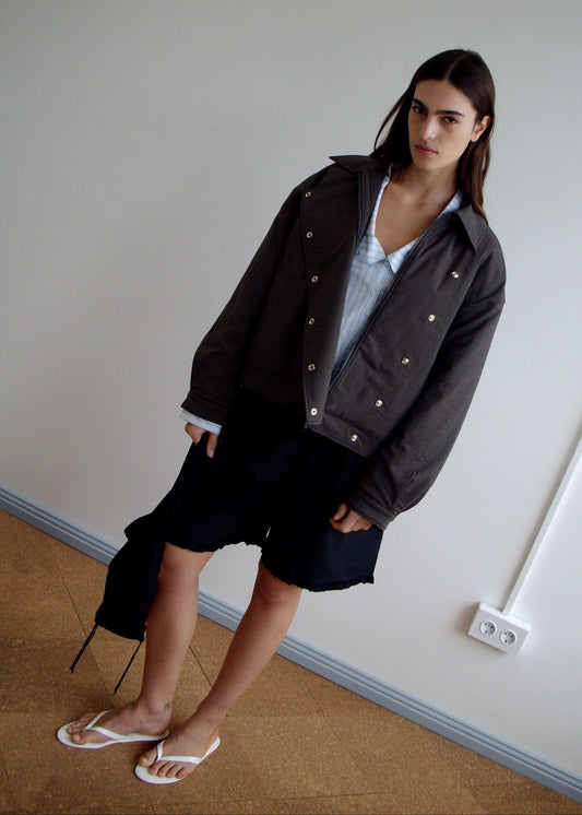 A woman stands indoors against a plain wall, wearing a dark oversized jacket, a light blue shirt, and Deiji Studios’ Flounce Short in black—one side features an asymmetrical, longer flounce hem. She finishes the look with white flip-flops.