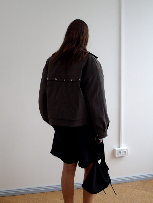 A person with long brown hair stands facing a white wall, wearing the Lapel Jacket - Brown Stripe from Deiji Studios and a black skirt with an uneven hem, holding a black bag in their right hand.