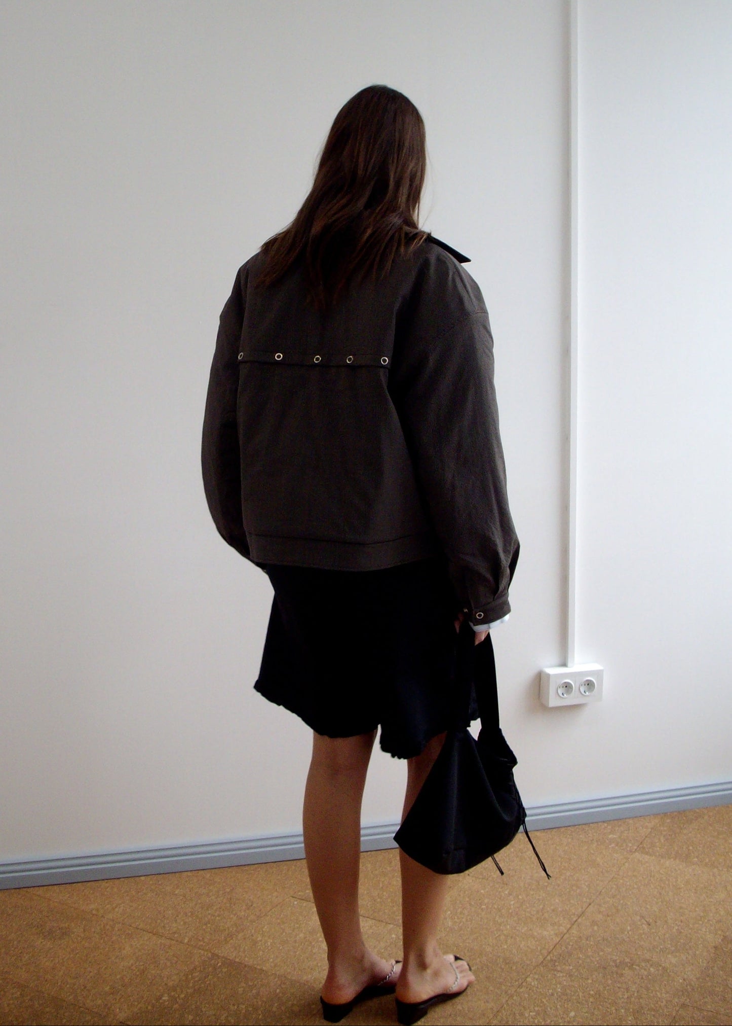 A person with long brown hair stands indoors facing away, wearing a dark oversized jacket, Flounce Short - Black, sandals, and holding a black bag. The plain white wall in the background has a visible power outlet.