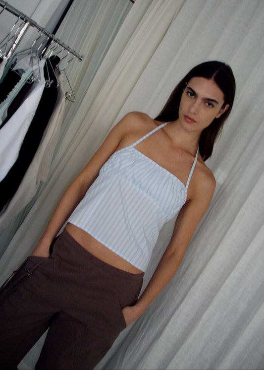 A woman with long dark hair and a neutral expression stands indoors, wearing the Strap Top - Mineral Stripe and brown pants. She has one hand in her pocket. Behind her are hanging clothes, a mirror, and white curtains.